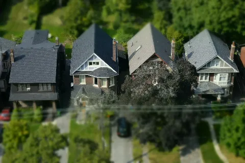 Low aerial drone view across the rooftops of four detached homes on a tree lined street, showing shingle condition, ridgelines, and chimneys. This is the vantage point a drone roof inspection uses to assess a roof without a ladder.