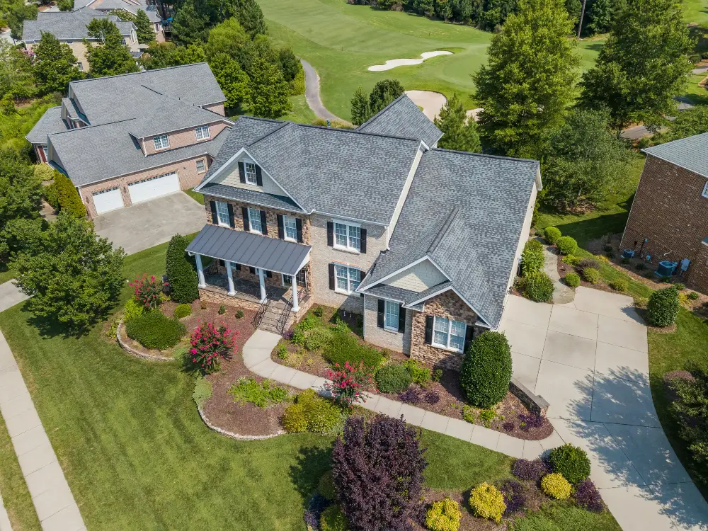 Low aerial view of a two storey brick executive home with landscaped gardens, a curved driveway, and a golf course visible beyond the treeline. The angle and framing are the kind a certified drone photographer plans in advance rather than captures by chance.
