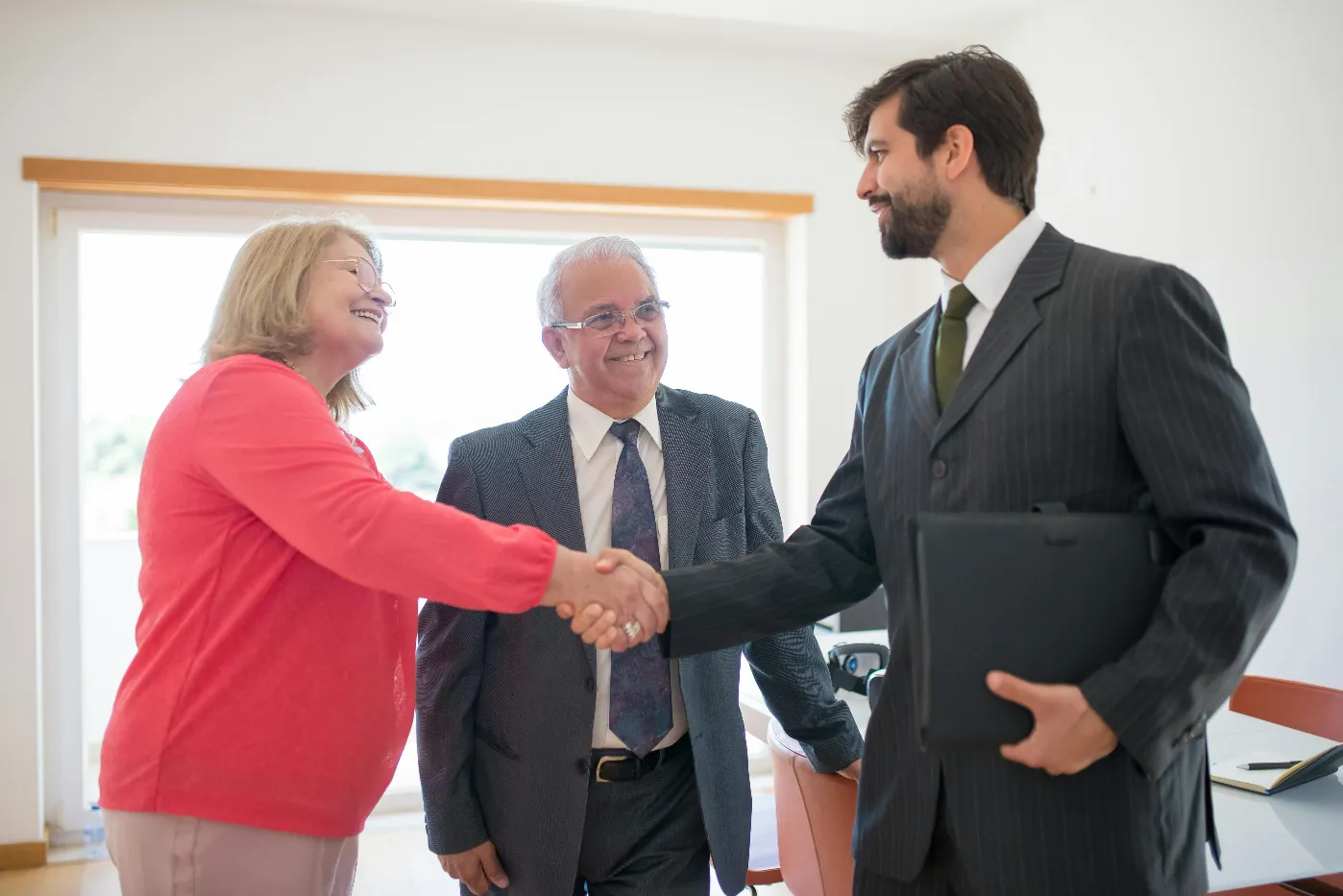 A real estate agent in a suit shakes hands with a smiling couple in a bright room after a listing presentation. Winning the listing often comes down to the marketing plan the agent presents.
